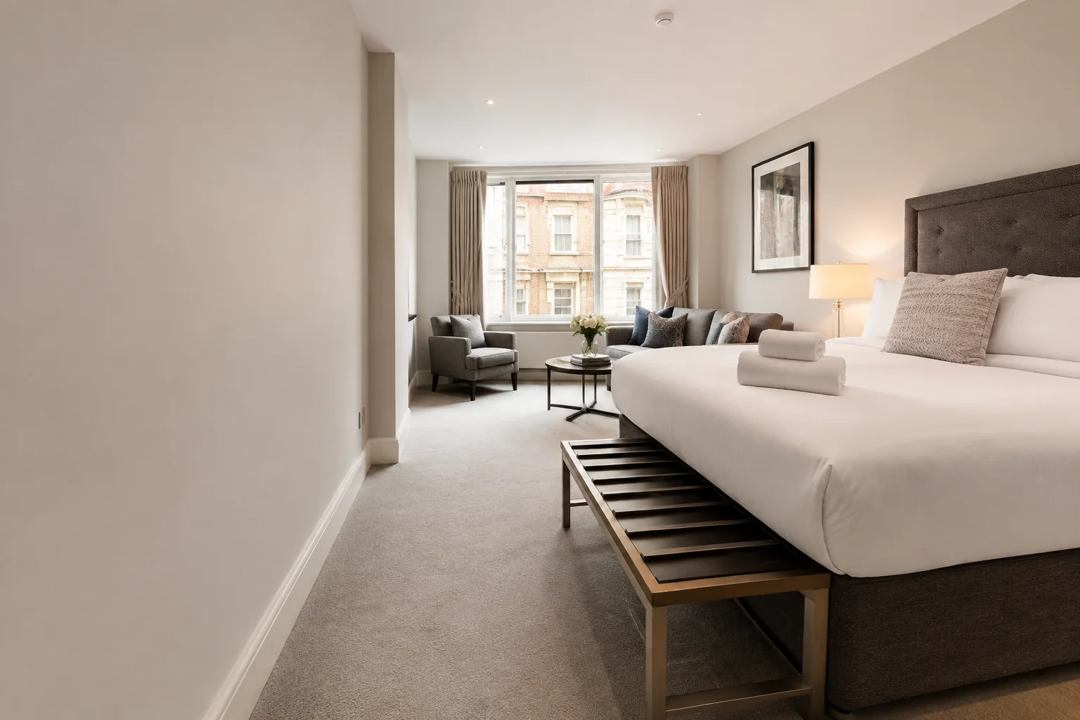 Prepared apartment bedroom with white linen, folded towels and a seating area